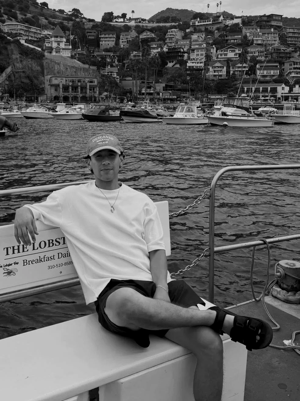 Joshua Galvan sitting on a boat in a Catalina Island harbor, white tee and Descanso Beach Club cap